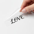 Peeling the transparent backing off a black Love UV transfer sticker to show the die-cut finish without plastic edges.