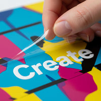 Hand peeling clear transfer film from a white UV sticker reading Create on a colorful background.